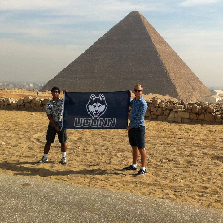 students hold a flag with the UConn Logo in front of a pyramid in Egypt