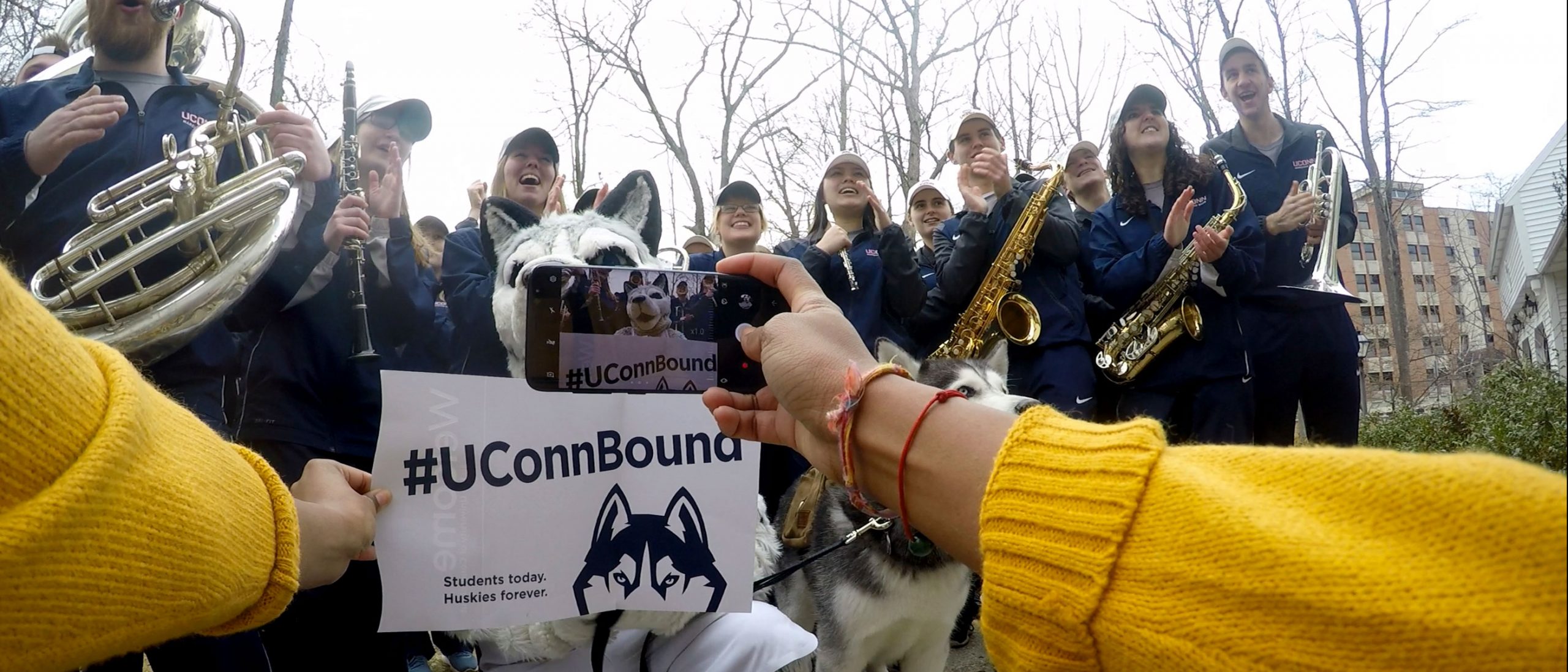 a cellphone captures a joyful moment of a UConn Bound acceptance letter given to Jonathan the husky while surrounded by a band