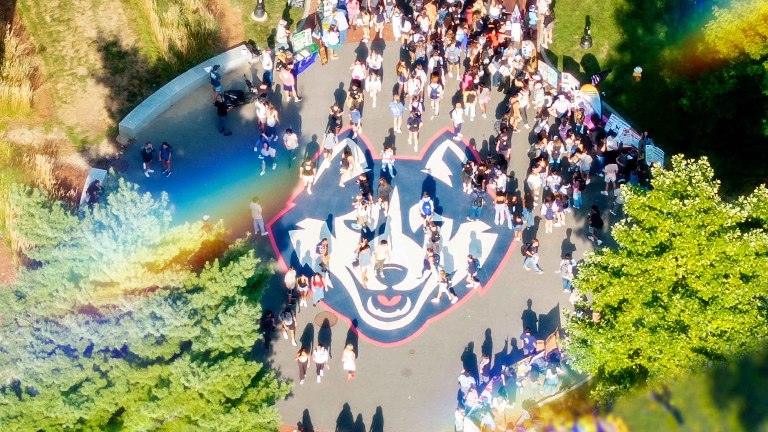a prism of rainbow light filters over Fairfield way. Hundres of students cross the pathway that features the UConn Husky head logo.
